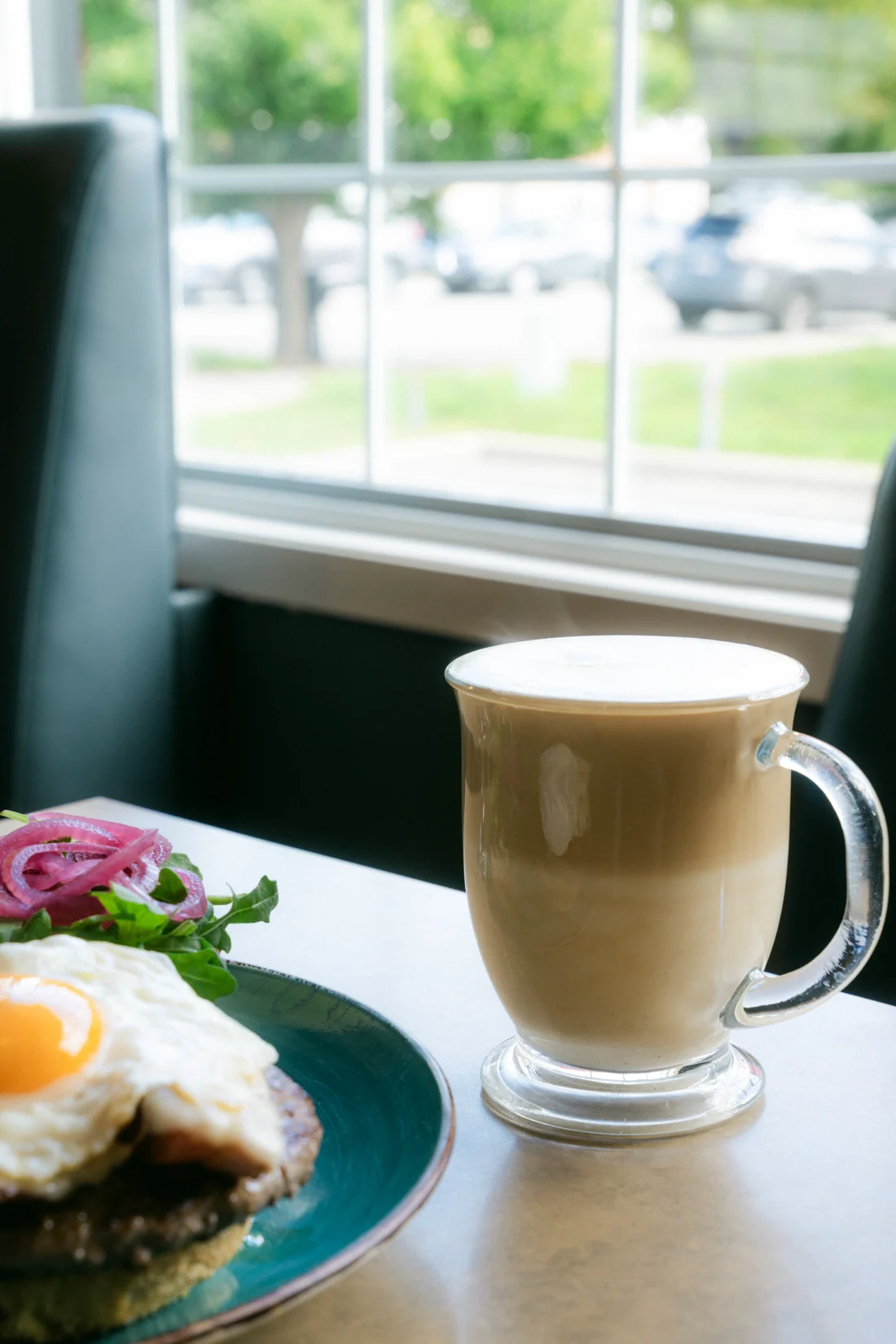 A latte in a glass mug by a diner window, a plate in the foreground