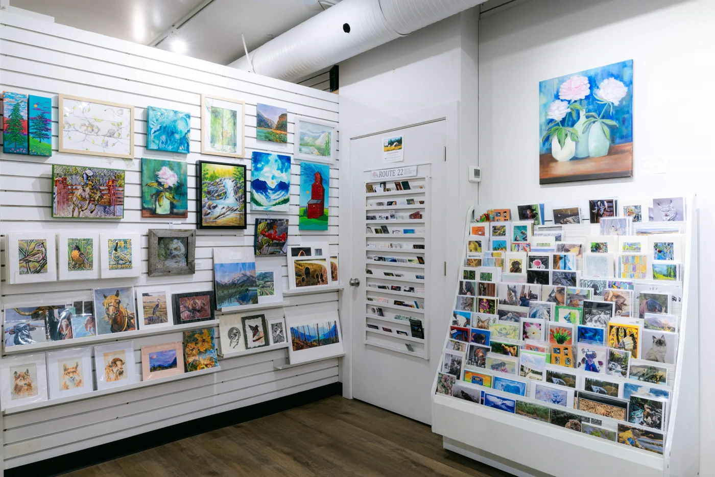 Small paintings hung close together on a gallery wall beside a rack of art cards and prints