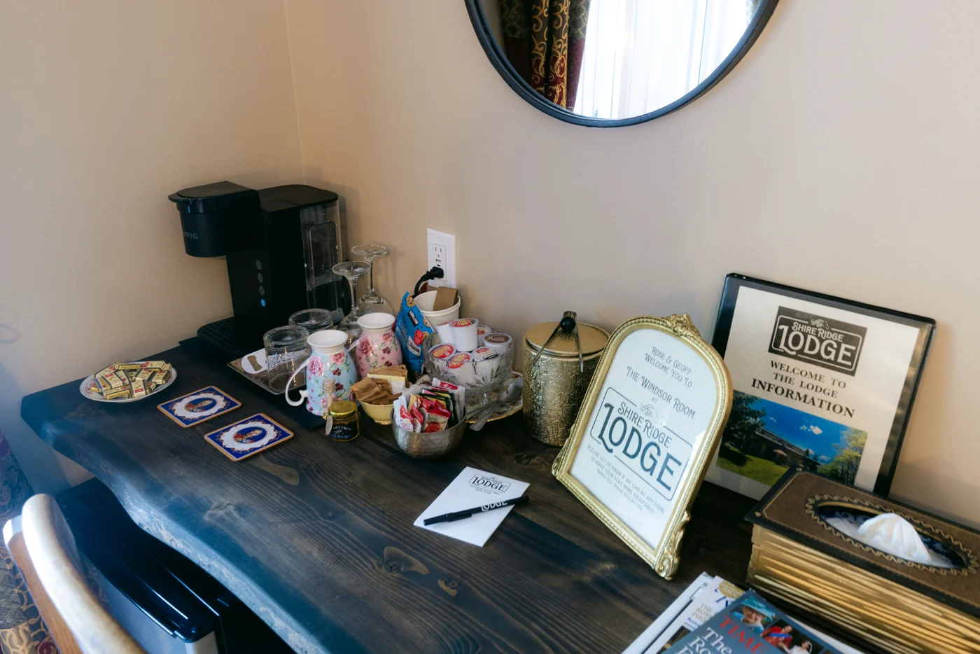A coffee maker, mugs and guest information on a lodge desk under a round mirror