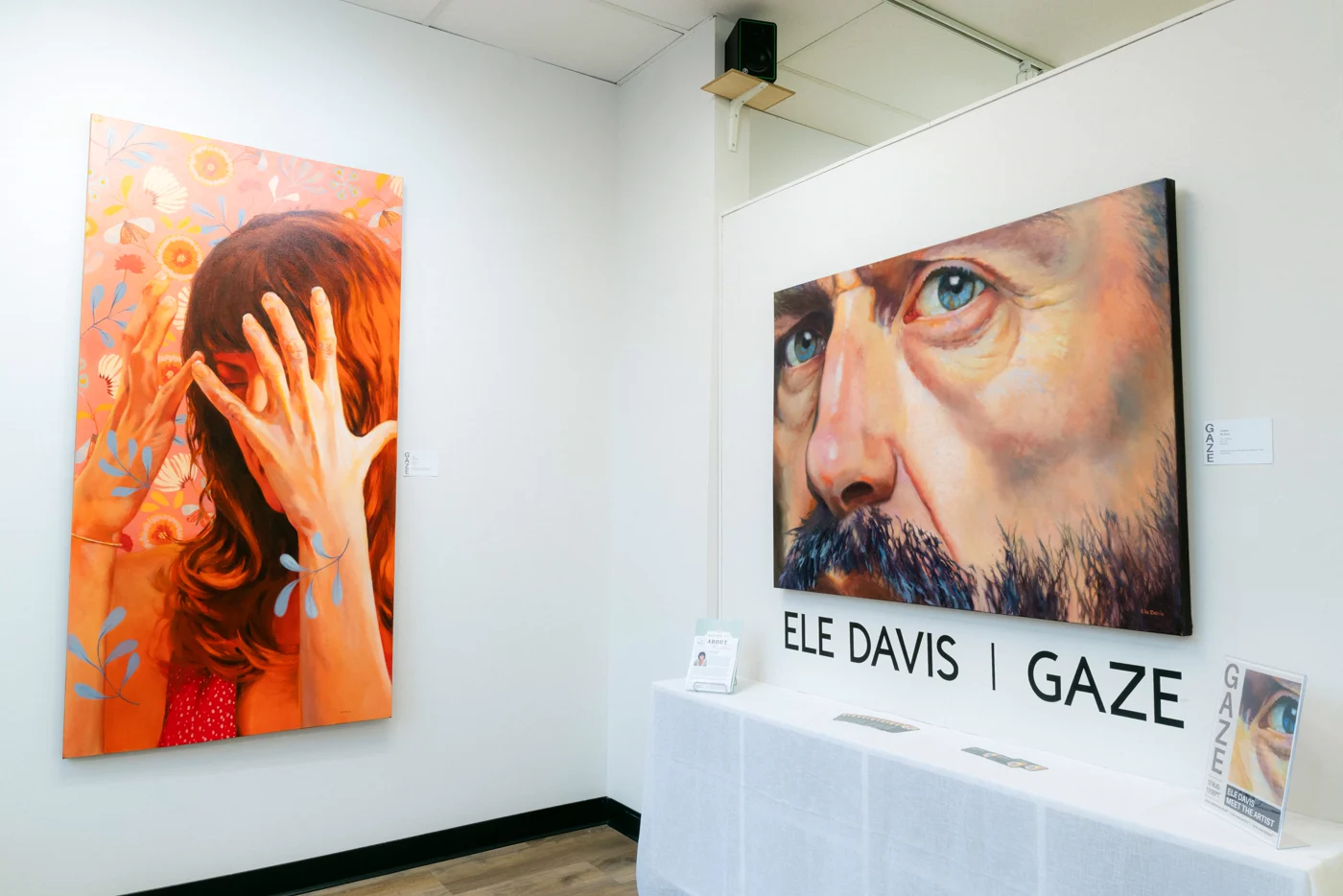 A gallery wall with a painted portrait of a woman covering her face and a large canvas of a man's eyes