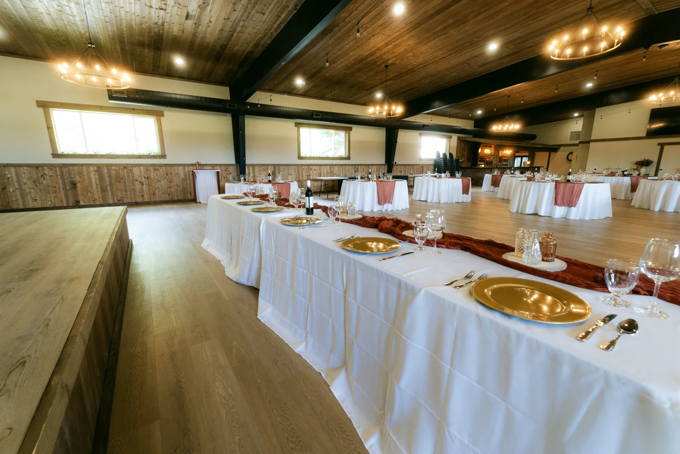 Long head tables set with gold chargers on a raised floor in a barn hall
