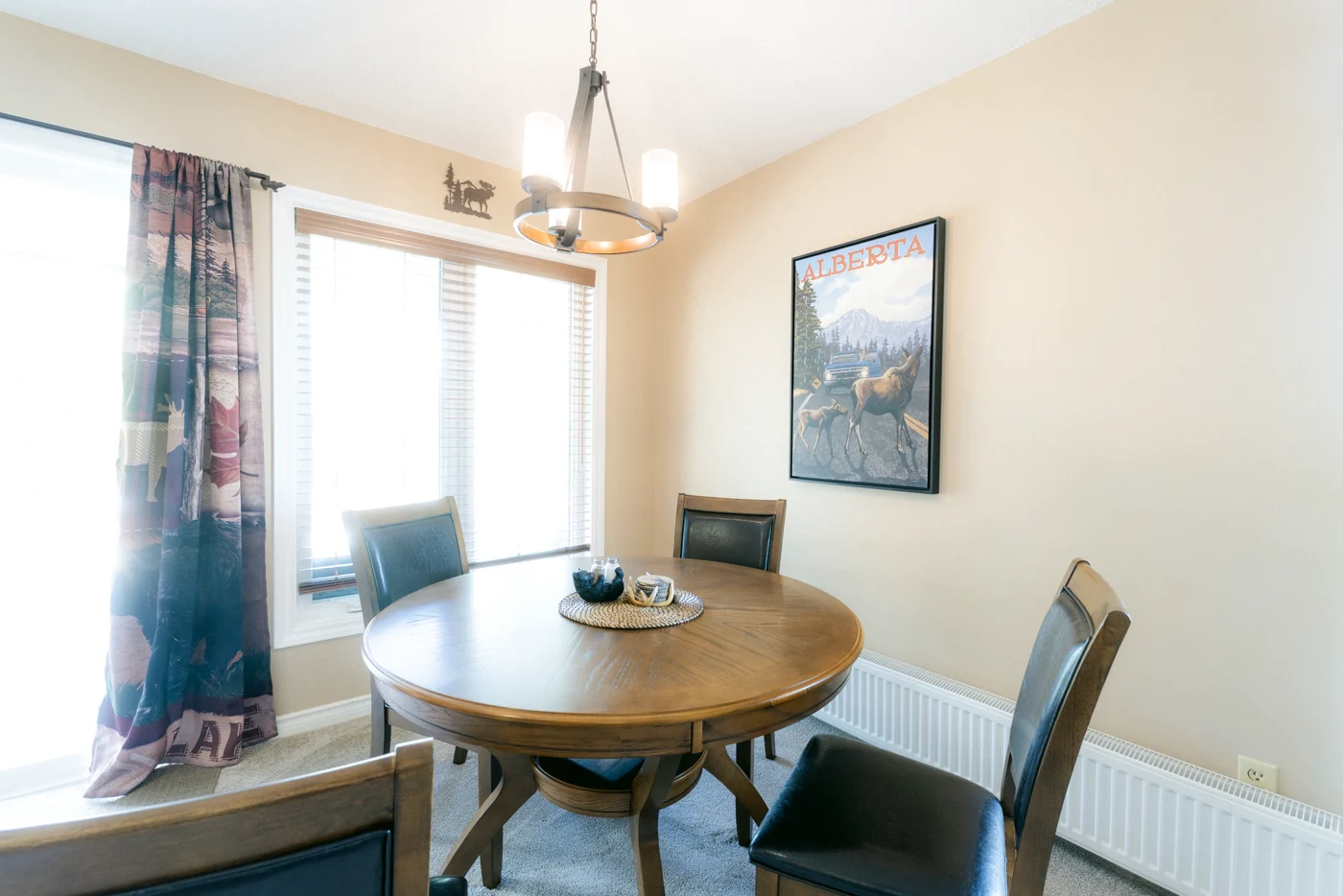 A round wooden dining table by a window under a chandelier and a vintage poster