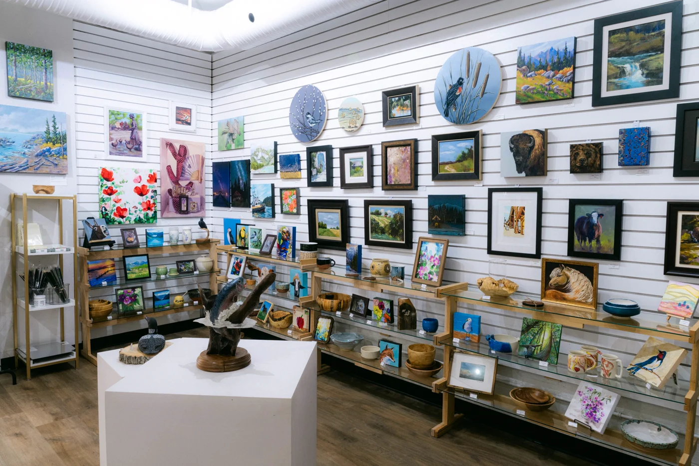 A white slatwall hung with framed paintings, a sculpture on a plinth and shelves of pottery in a gallery shop