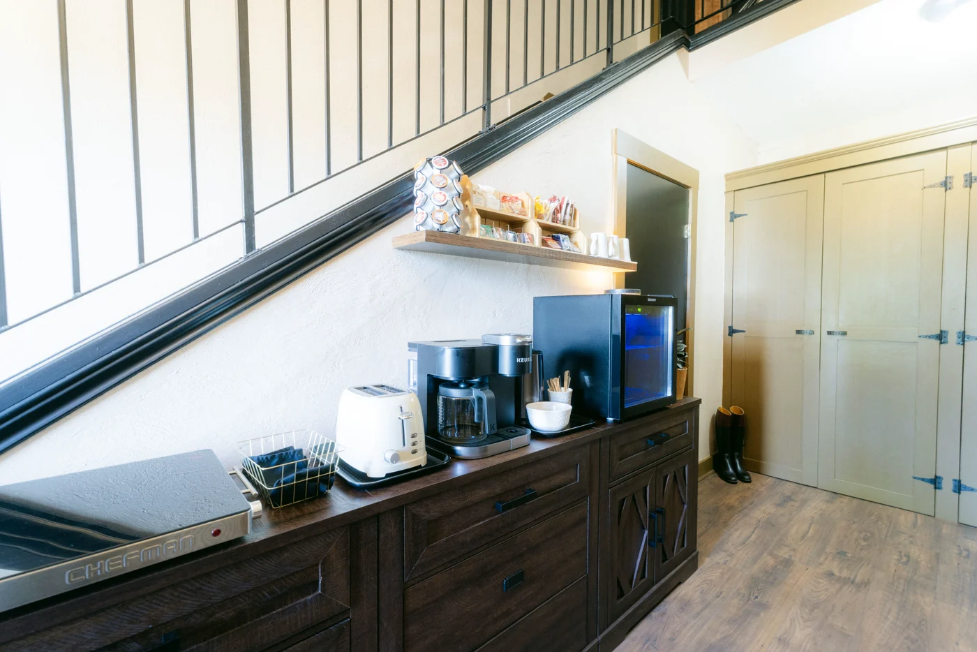A coffee station on a dark sideboard under a lodge staircase