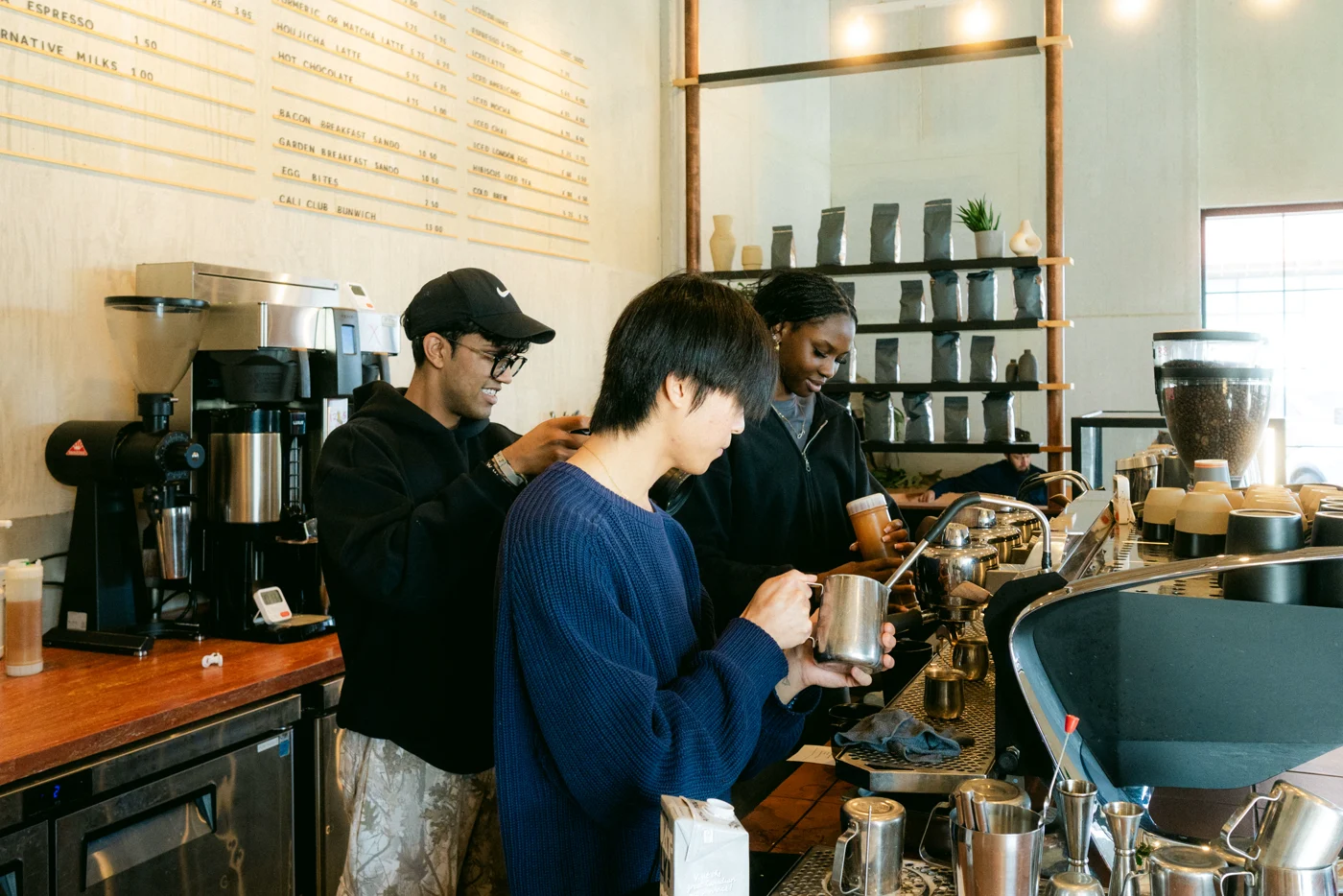 A photographer in a black cap and hoodie frames two baristas steaming milk at the espresso bar