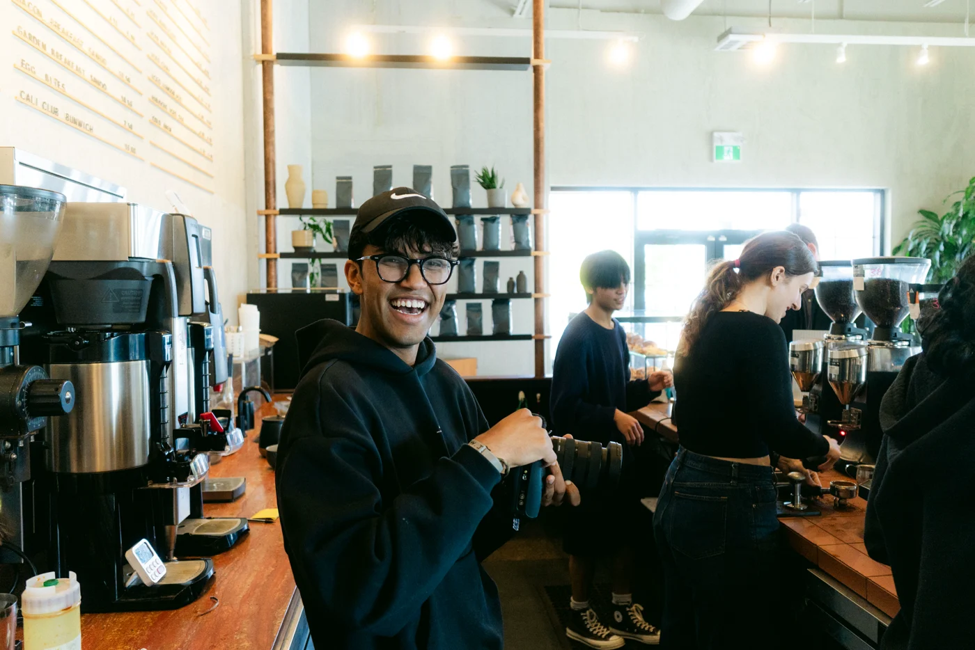 A photographer laughs with two baristas behind the bar of a café, camera in hand