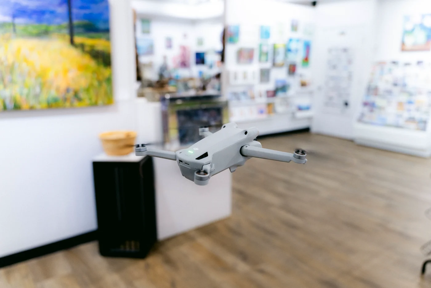 A small drone hovering at eye level in an art gallery, paintings and shelves blurred behind it