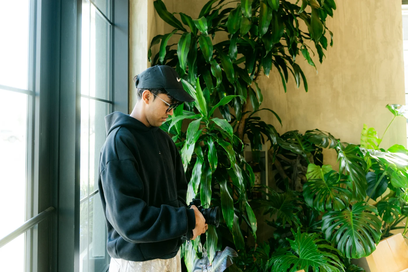 A crew member in a black cap checks his gear among tall plants at a café window