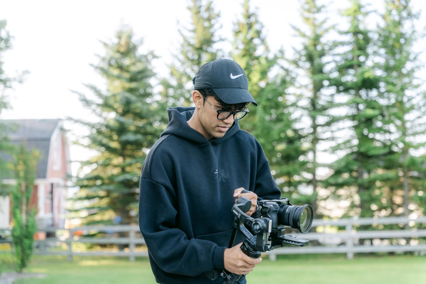 A camera operator with a gimbal rig on a lawn in front of spruce trees
