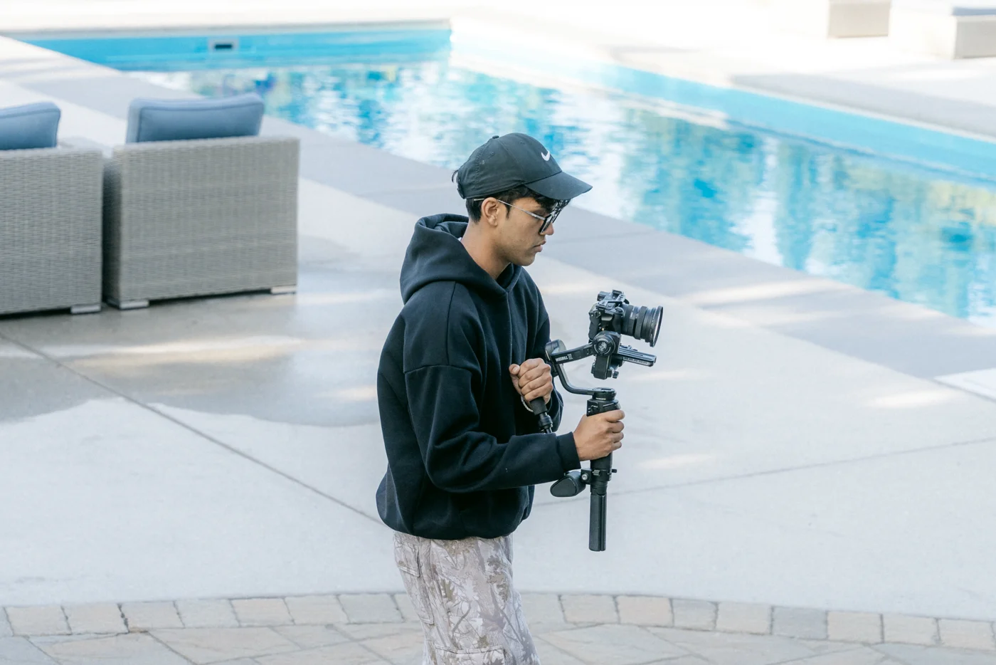 A camera operator carries a gimbal rig along the edge of a backyard pool