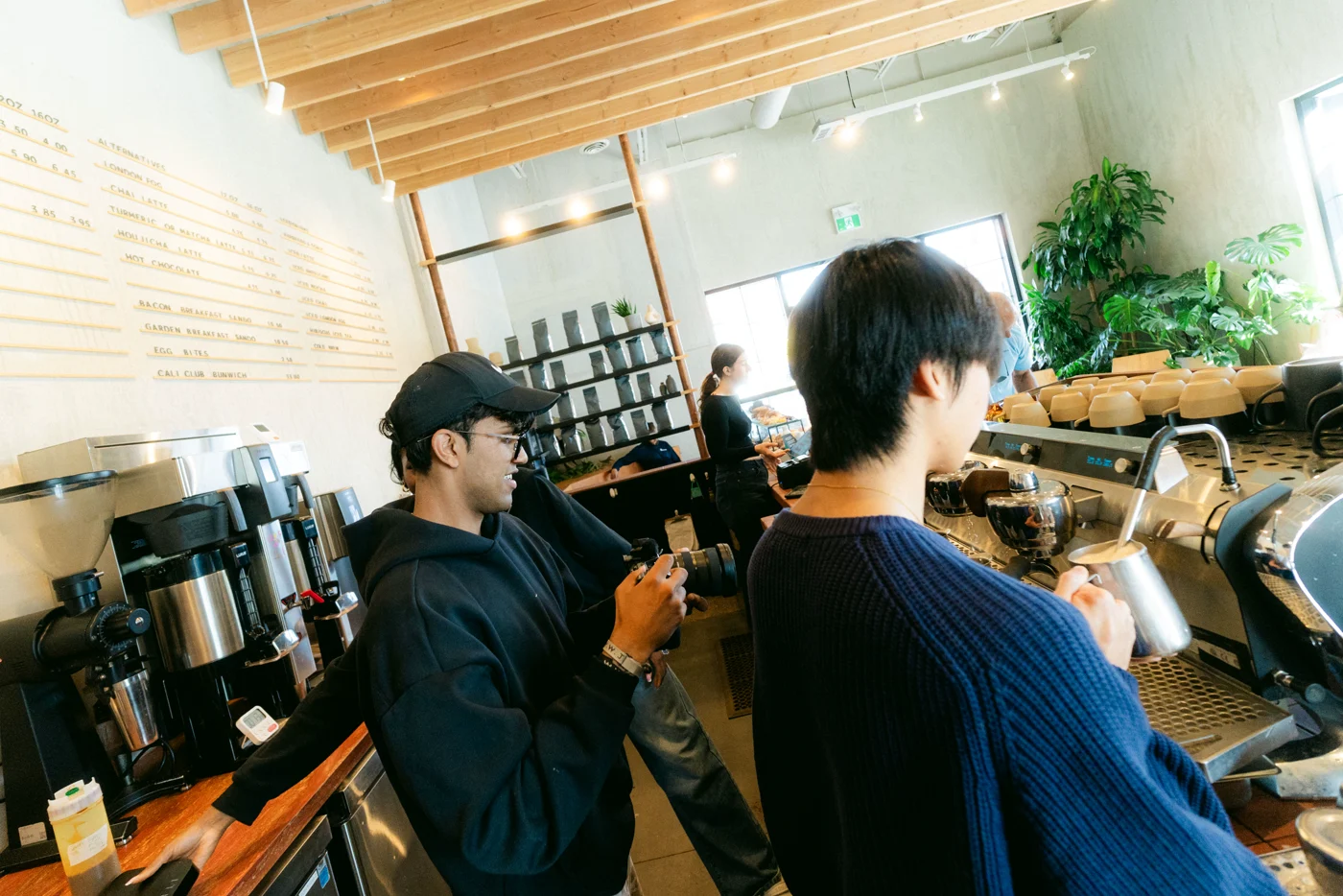 Seen from behind, a photographer lines up a shot of a barista at the espresso machine in a café