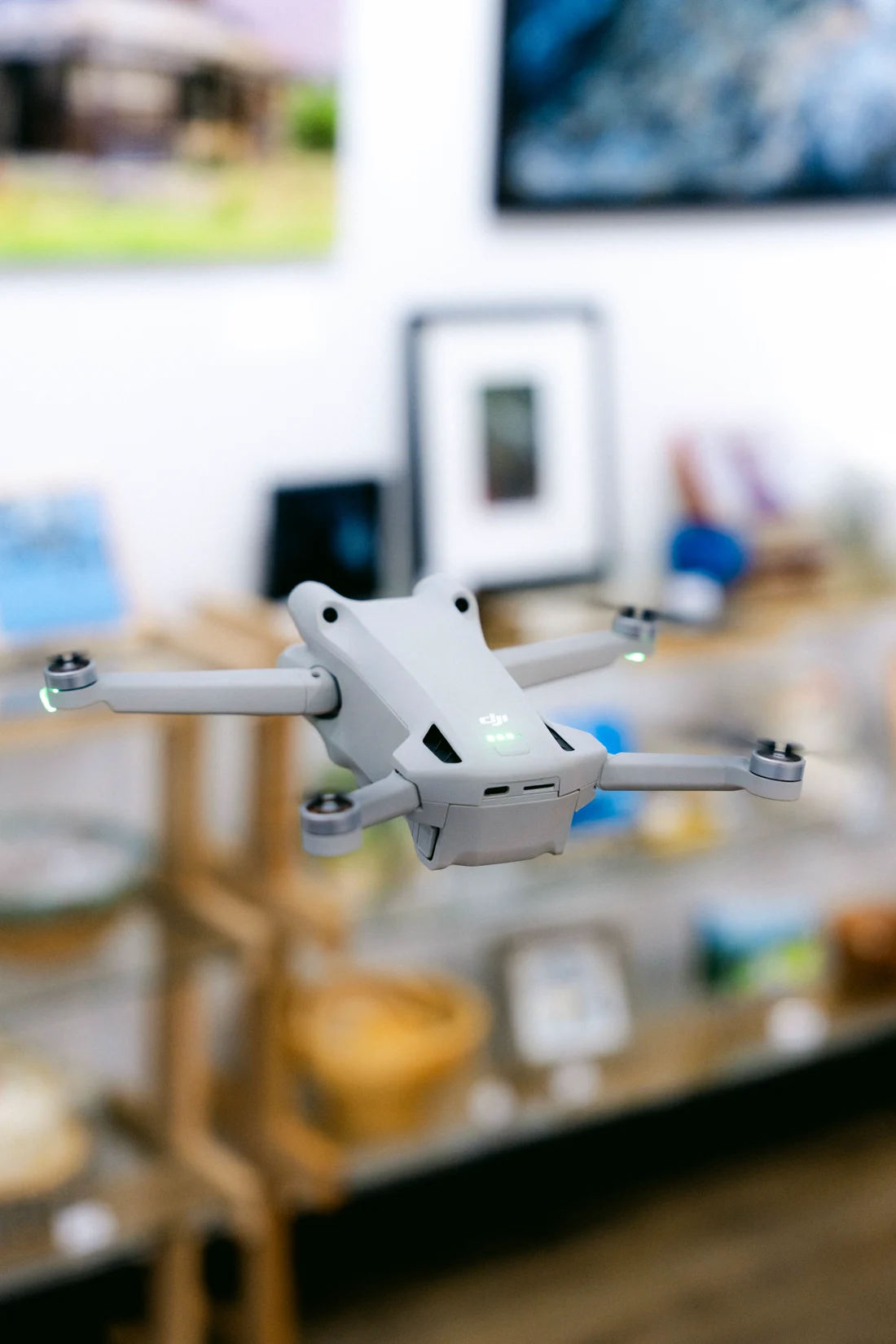 A small grey drone hovering inside a gallery shop, shelves of pottery and prints softly out of focus behind it