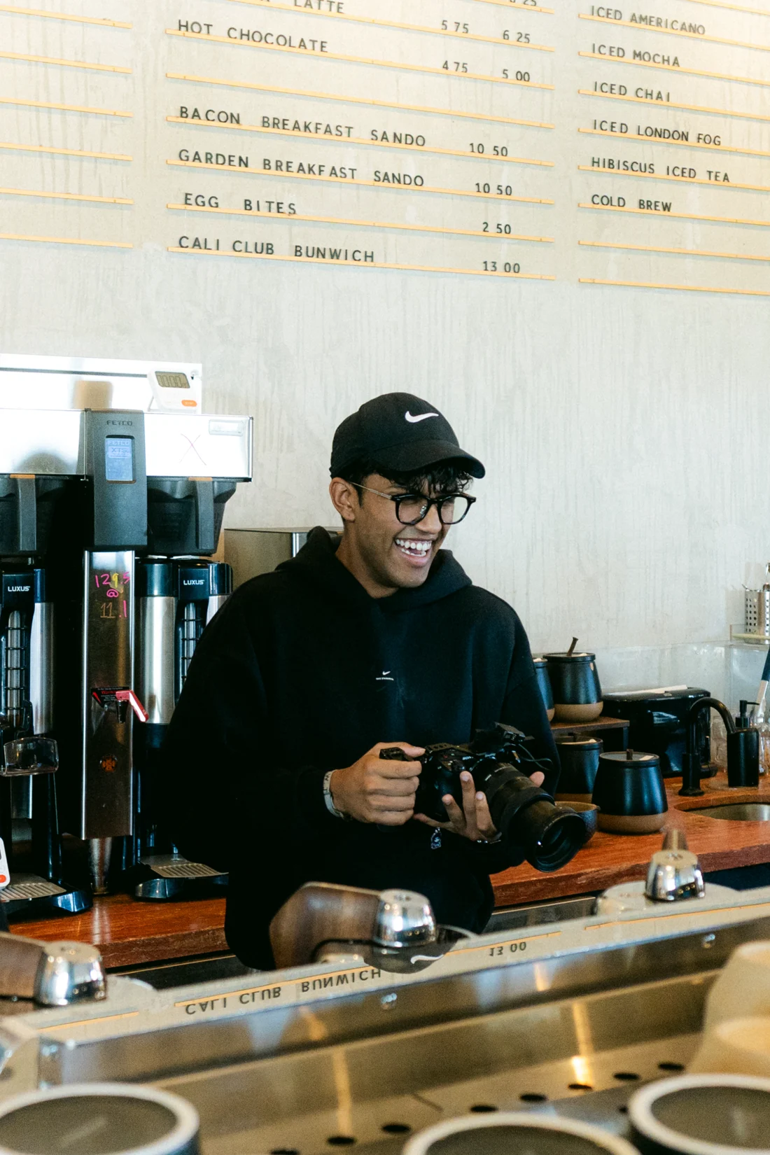 A photographer smiles with his camera behind the counter, a hand-lettered menu board on the wall above
