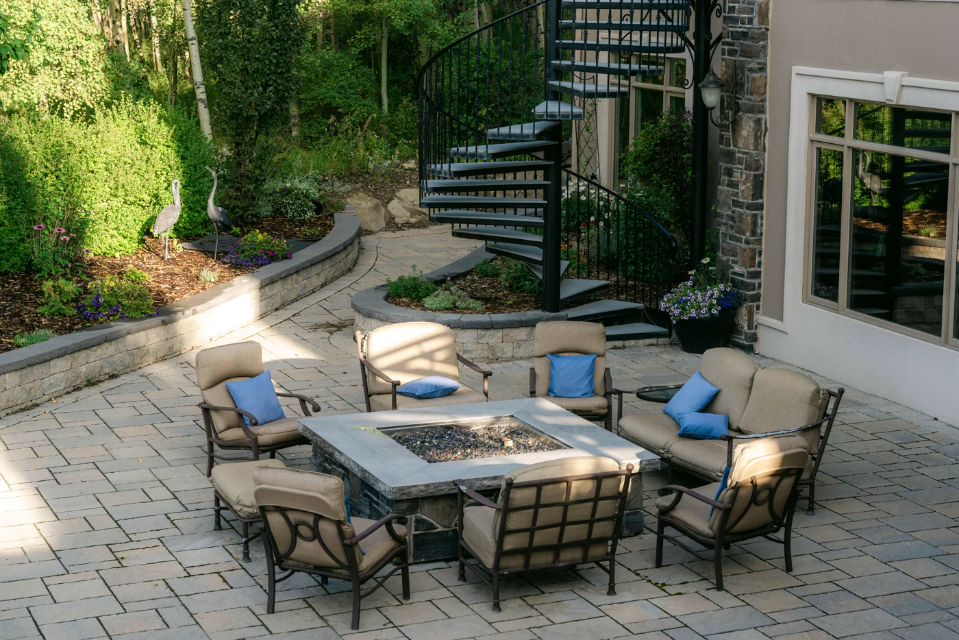 A patio with a stone fire table and armchairs below a spiral staircase