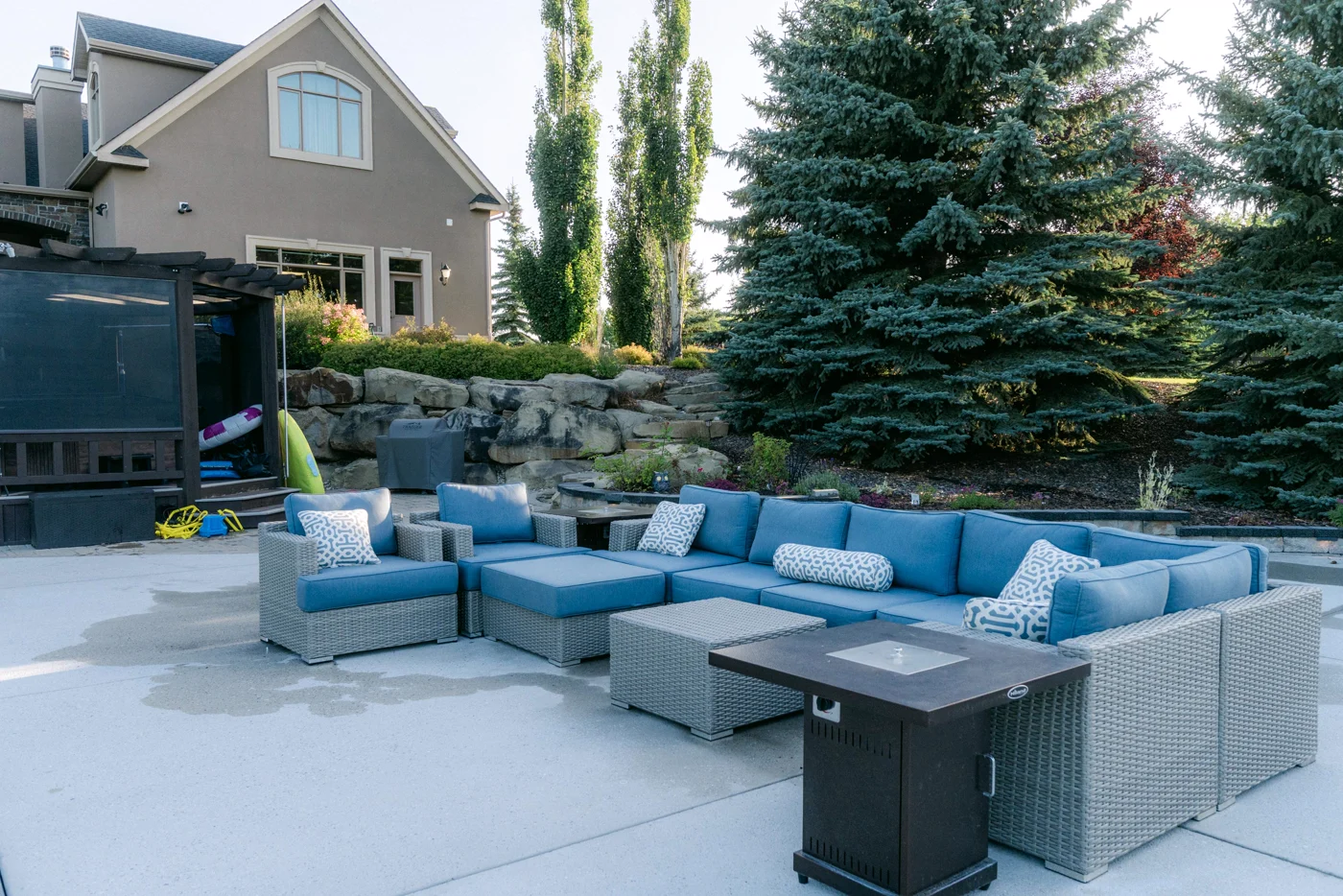 A grey sectional sofa with blue cushions around a fire table on a patio below spruce trees