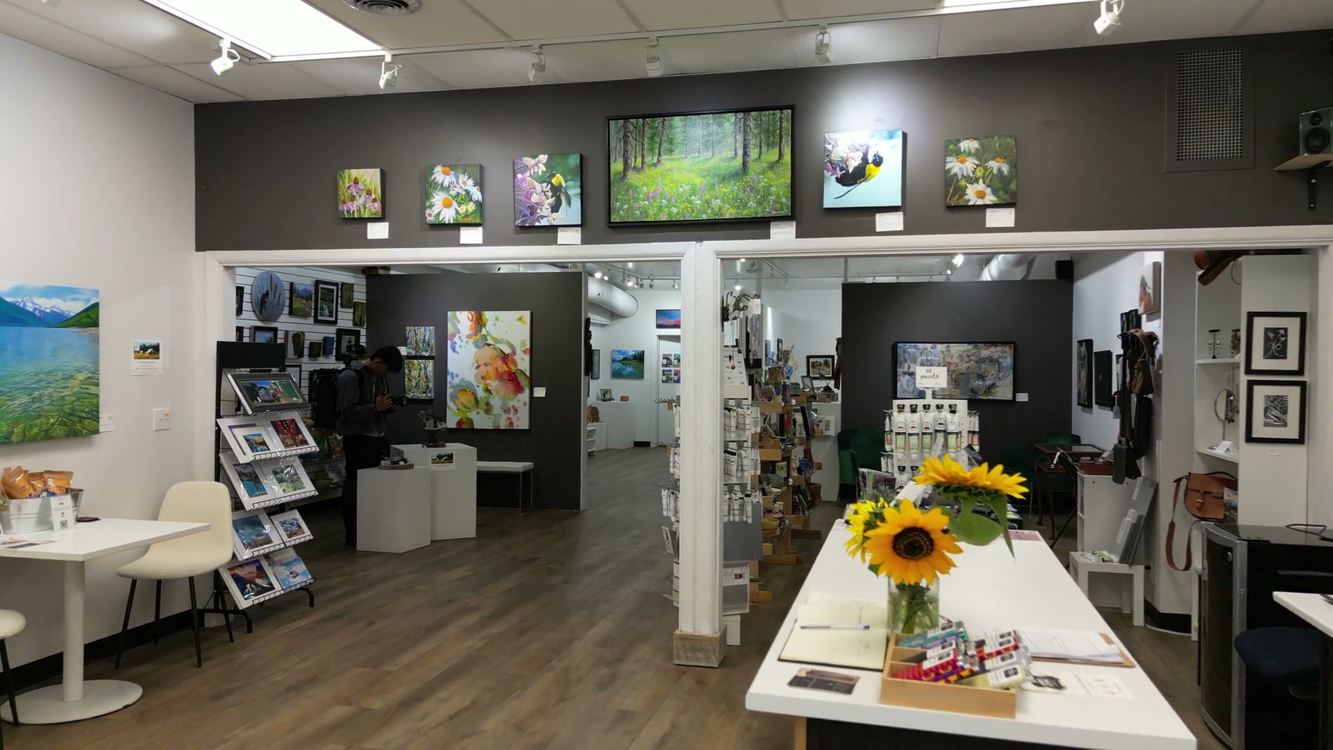 Drawing back from the gallery counter and its jar of sunflowers, through the opening into the far room