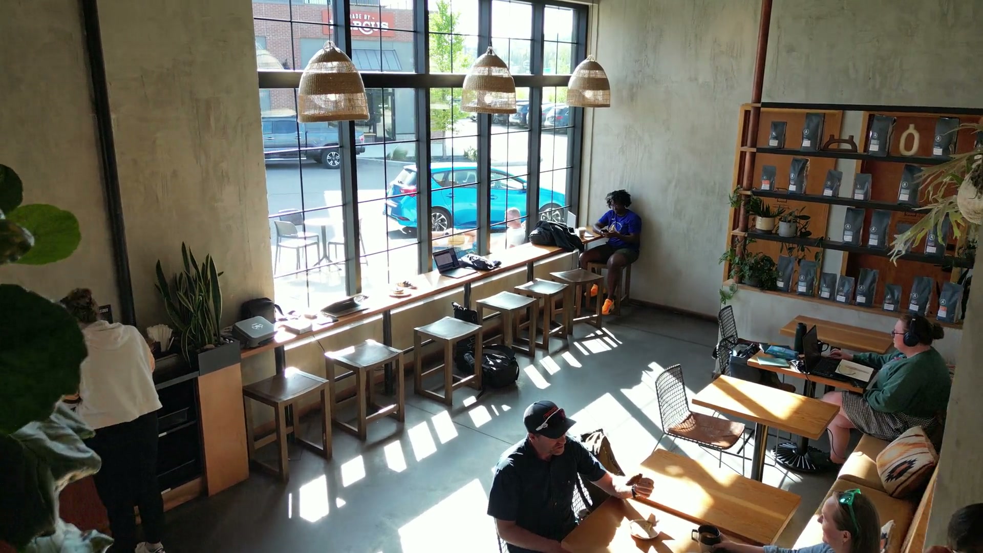 Inside at ceiling height, past a fiddle-leaf fig and the woven pendant shades, down the window counter where laptops are open on the sill