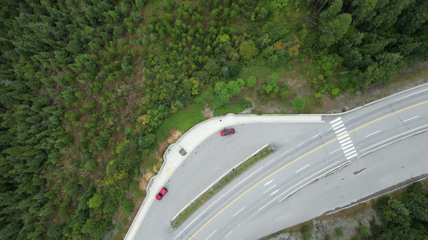 A highway junction near Radium Hot Springs from directly overhead: a triangular median with a guardrail, painted crosswalk bars, and cars parked in the pull-out beside it