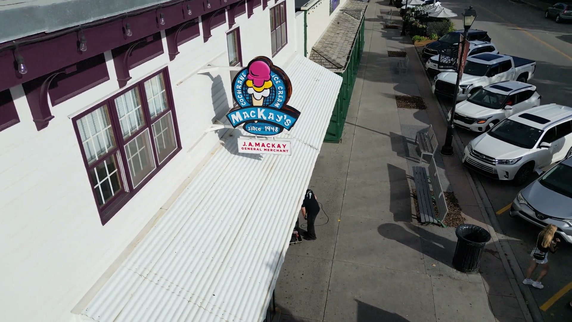 Descending past the shop's corrugated awning to the MacKay's roundel, pedestrians on the sidewalk below