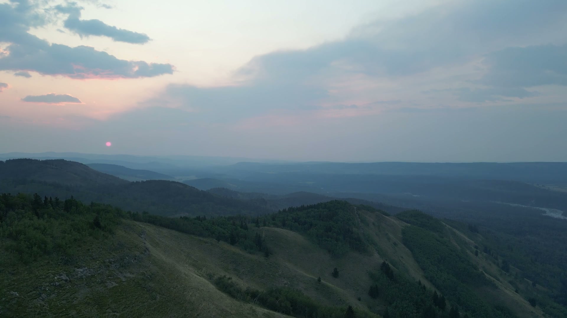 The sun as a pink-red disc just clear of the far ridges at sunrise, the drone drifting at half speed over layered forested ridges in the Alberta foothills