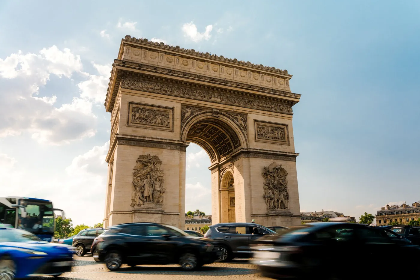 Traffic passing a triumphal stone arch under a bright cloudy sky