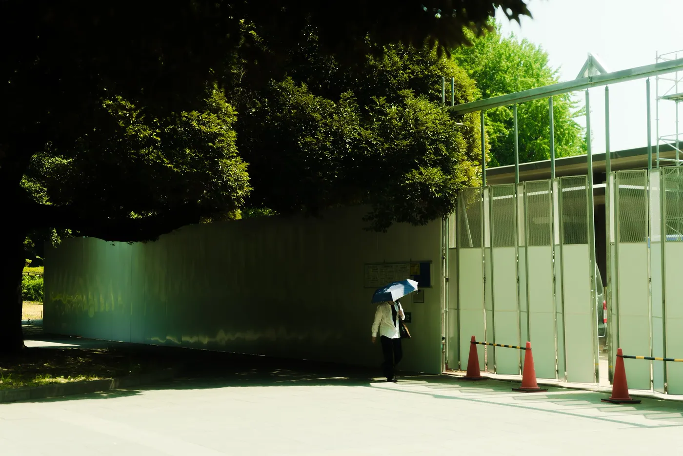 A lone figure with an umbrella beside a green glass structure under heavy trees