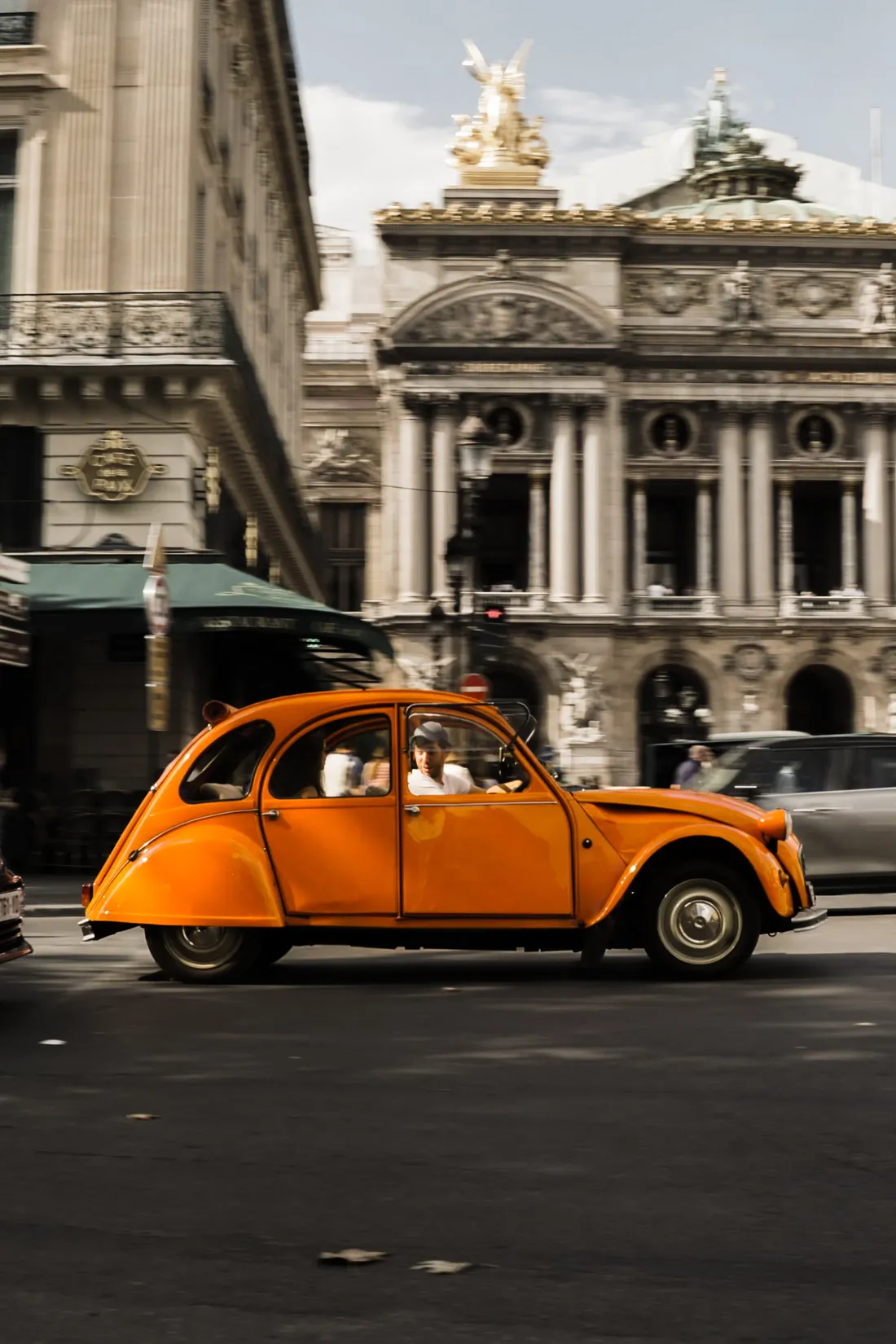 A small orange classic car passing an ornate opera-house facade