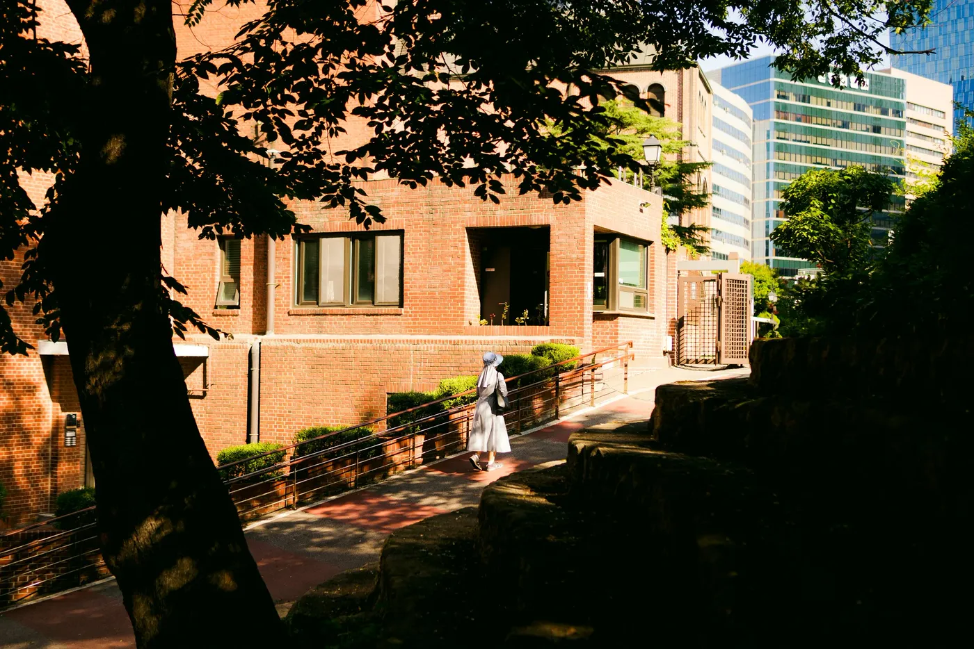 A woman in a white dress walking past a brick building, framed by dark foliage