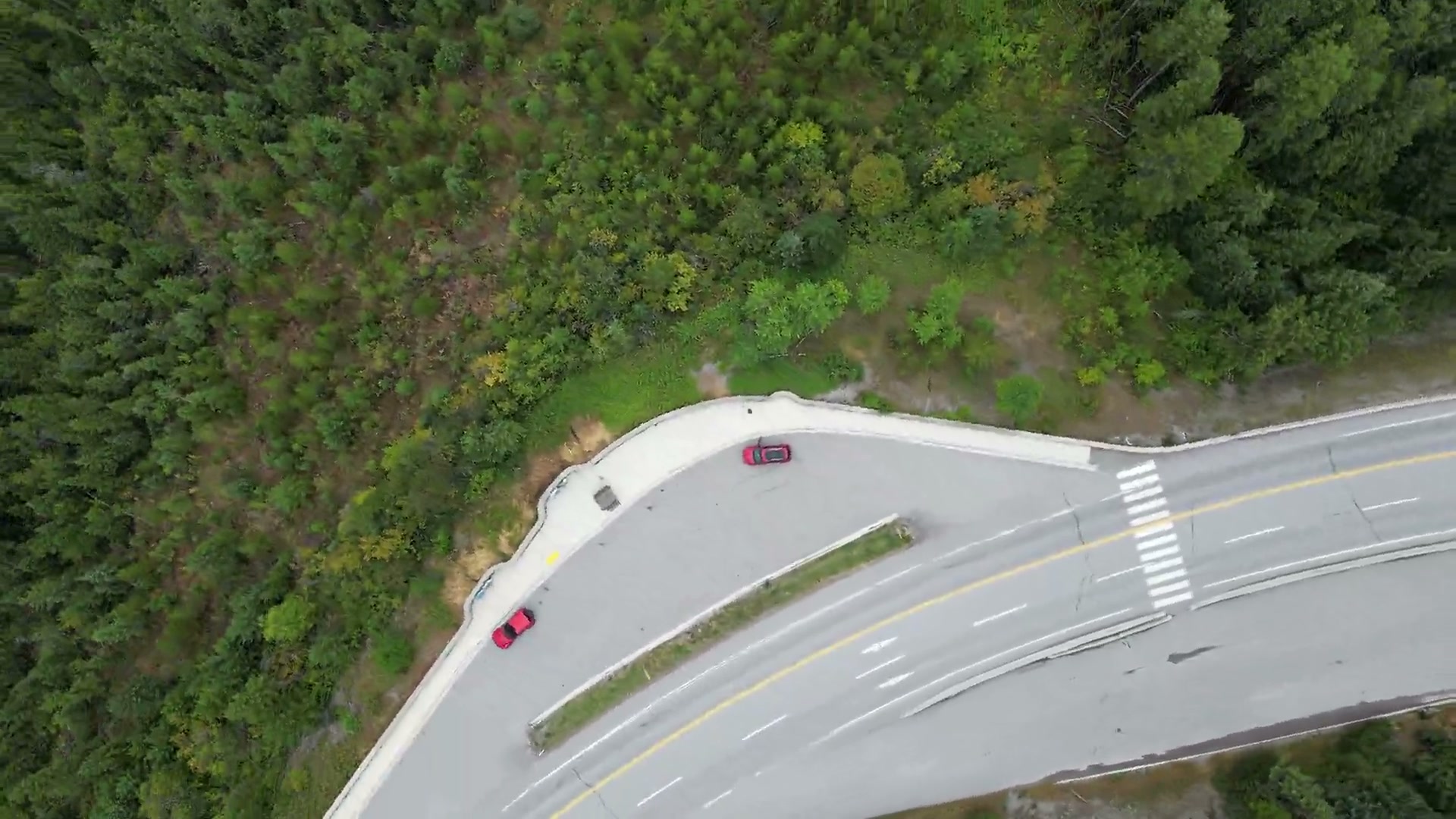 Directly above a highway junction in the forest near Radium Hot Springs: a triangular median with a guardrail, painted crosswalk bars, and a pull-out with cars parked in it