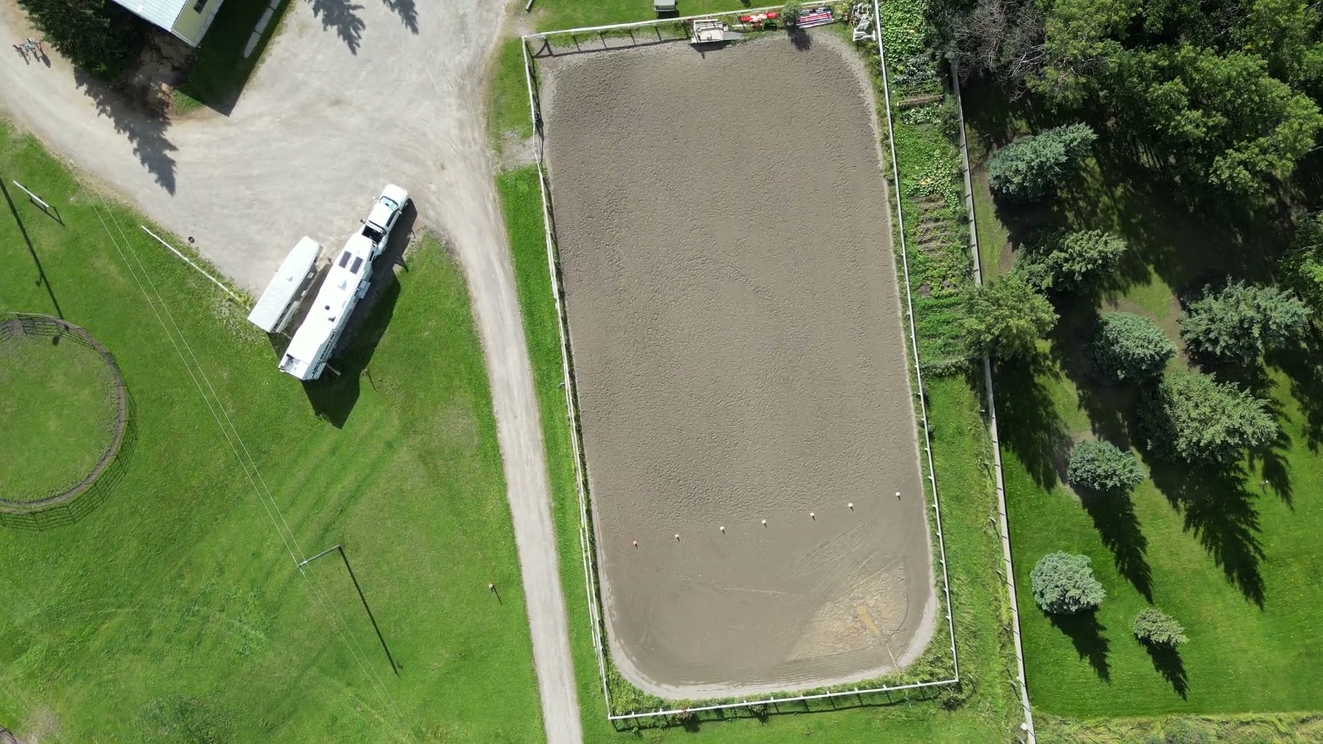 Straight down over a fenced sand riding ring at Shire Ridge Ranch near Bragg Creek, a pickup and a horse trailer parked beside it, a round pen and a row of trees at its edges