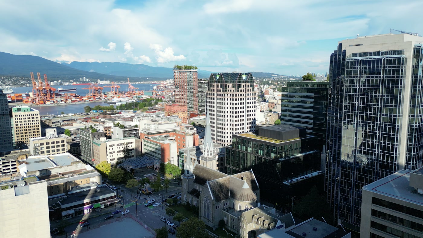 Downtown Vancouver from above: Holy Rosary Cathedral's two unequal towers at Richards and Dunsmuir, office towers around it, the container cranes across the harbour and the North Shore mountains under cloud