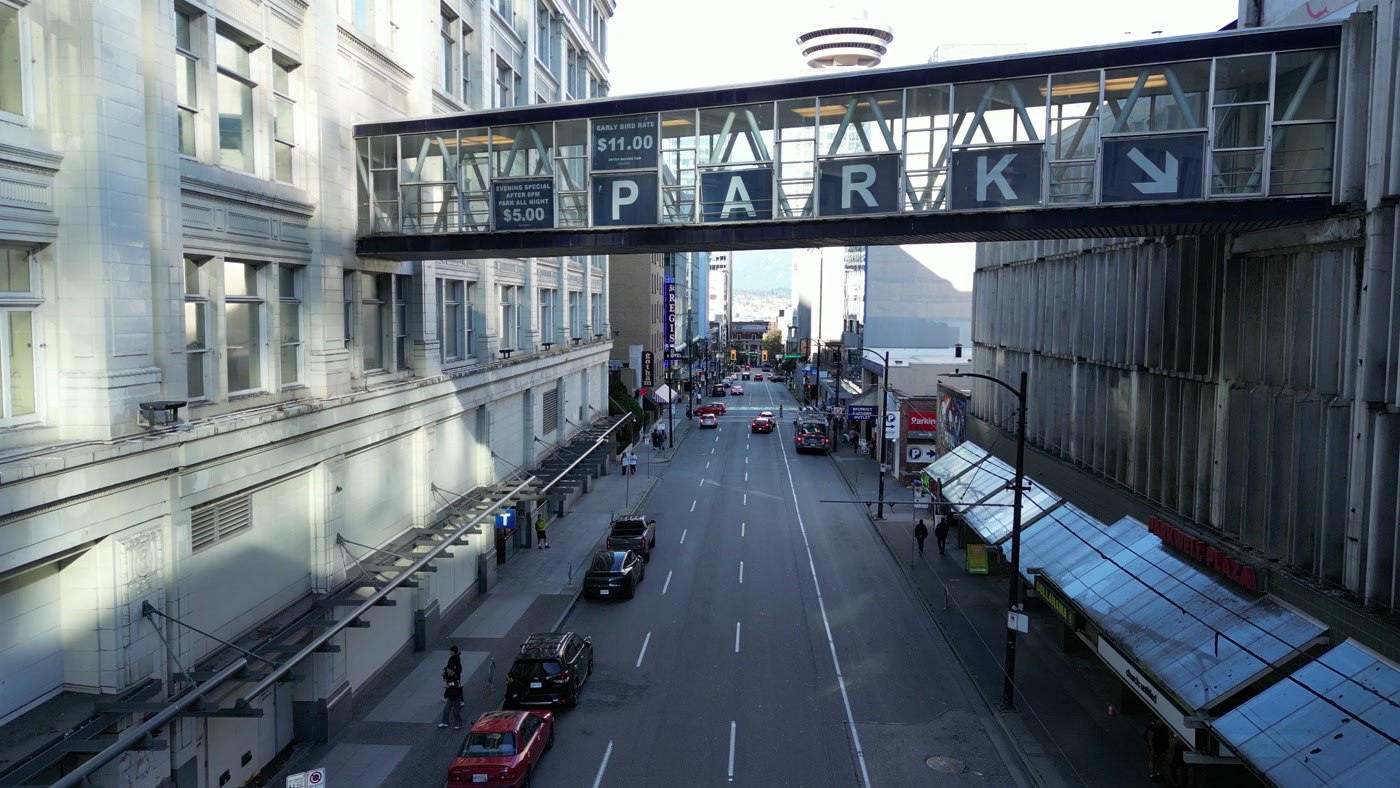 A glass skybridge across Seymour Street in Vancouver spelling P A R K with an arrow, its signs reading EARLY BIRD RATE $11.00 and EVENING SPECIAL AFTER 6PM PARK ALL NIGHT $5.00