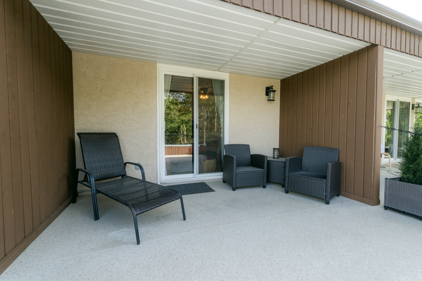 A covered balcony alcove at Shire Ridge Lodge near Bragg Creek: a reclining lounger and two dark armchairs around a small table, a wall lantern and a sliding glass door behind