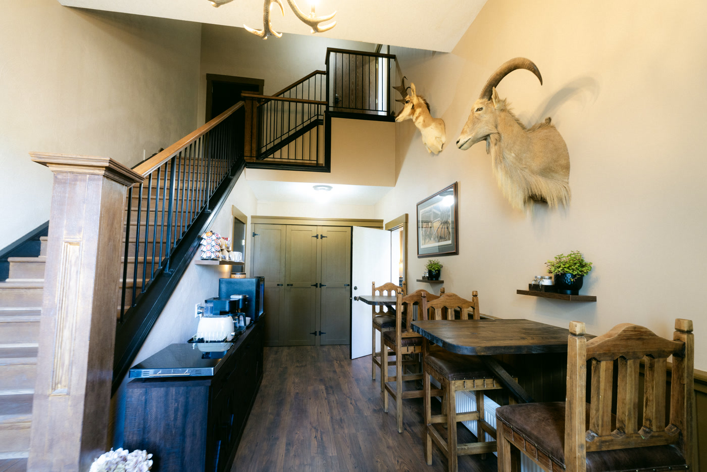 The breakfast area of Shire Ridge Lodge near Bragg Creek: the staircase with black railings, a coffee station, tables with carved wooden chairs, mounted heads and a framed print