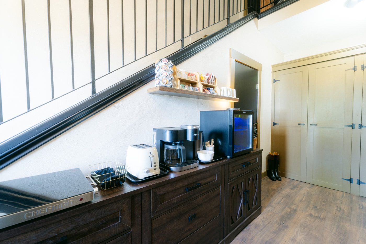 The coffee station tucked under the staircase at Shire Ridge Lodge near Bragg Creek: a dark sideboard with a toaster, a coffee maker, a small fridge and a shelf of mugs and treats