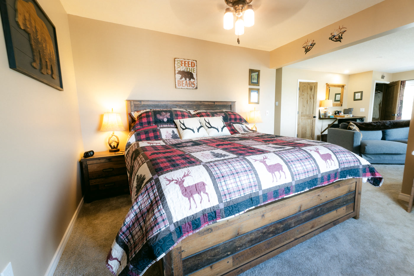 The Elk Suite's king bed head-on at Shire Ridge Lodge near Bragg Creek: country plaids with deer patches, antler pillows, two lamps, and the sitting room seen through the doorway