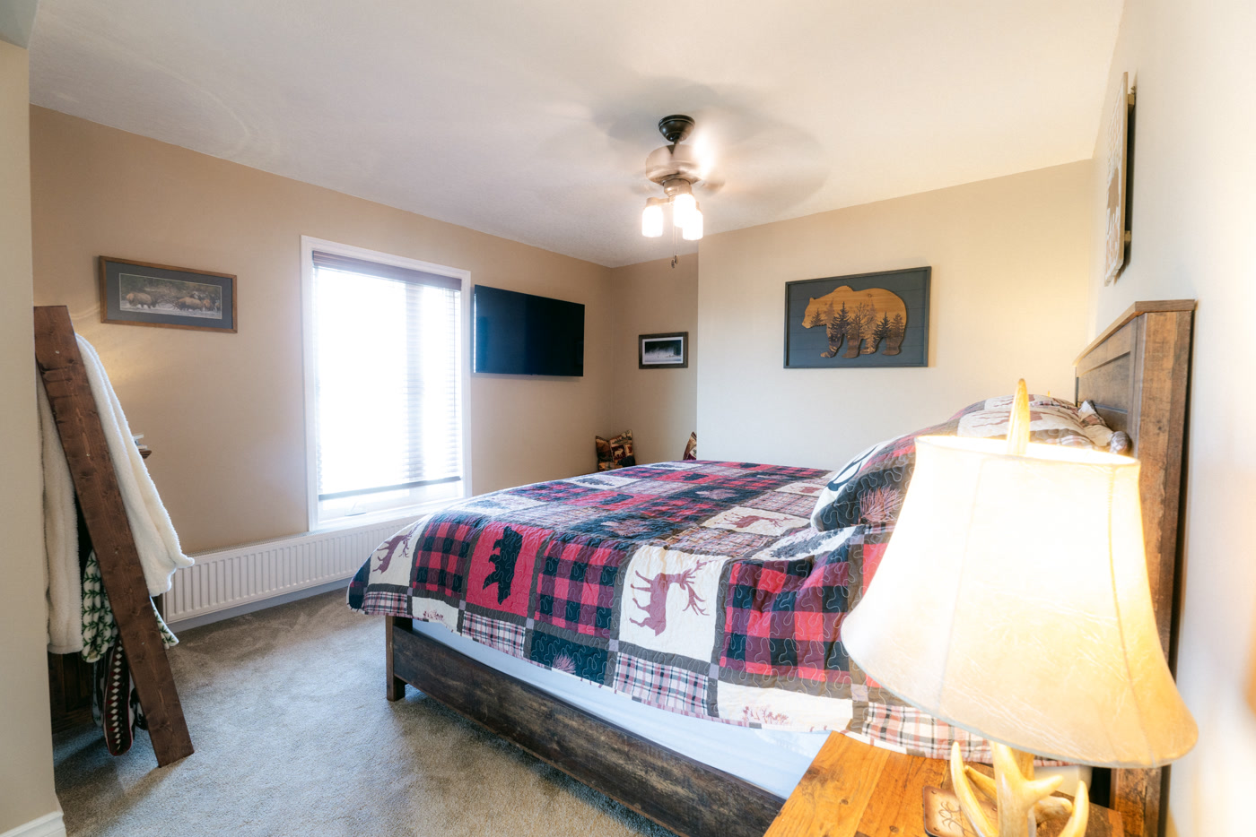 The Elk Suite's king bed at Shire Ridge Lodge near Bragg Creek under a red-and-black plaid quilt with deer patches, a lit lamp in front, a blanket ladder and a bison print behind