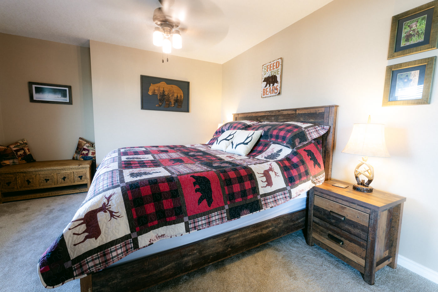 The Elk Suite's bed at Shire Ridge Lodge near Bragg Creek: a patchwork plaid quilt of deer and bears, antler-print pillows, a rustic nightstand with a lamp and framed prints
