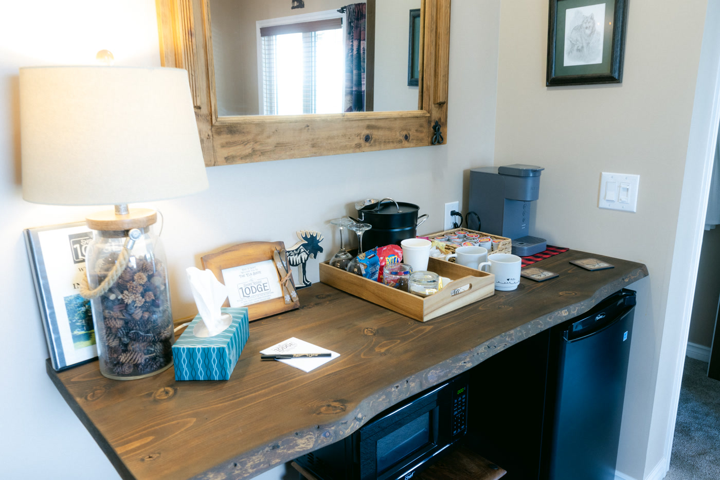 The live-edge wooden desk in the Elk Suite at Shire Ridge Lodge near Bragg Creek: a coffee machine, a tray of snacks, a jar of pinecones, a lamp and a timber-framed mirror