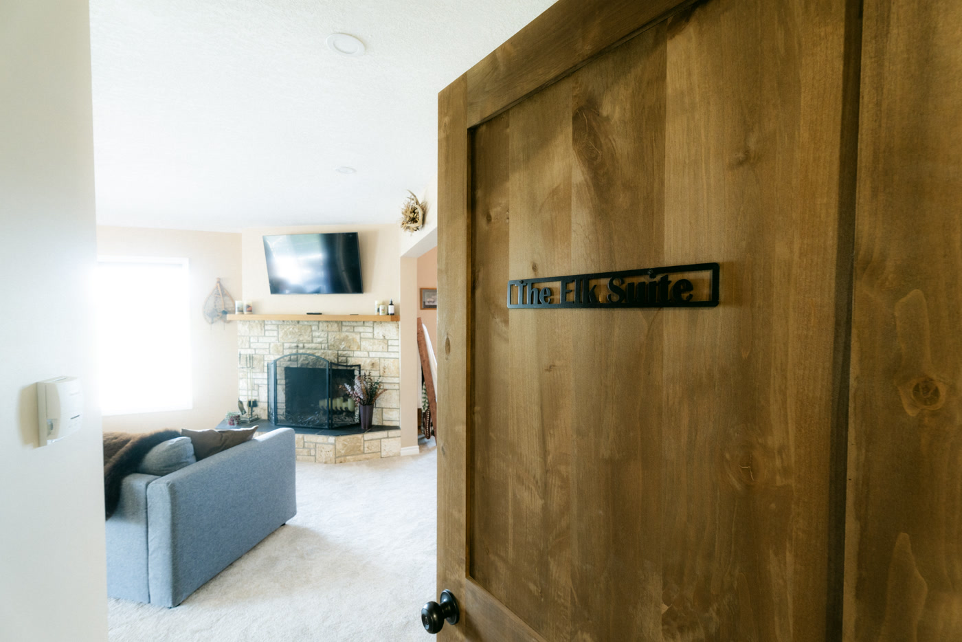 The black metal sign reading The Elk Suite on a knotty wooden door at Shire Ridge Lodge near Bragg Creek, the sitting room's stone fireplace and grey sofa visible beyond it