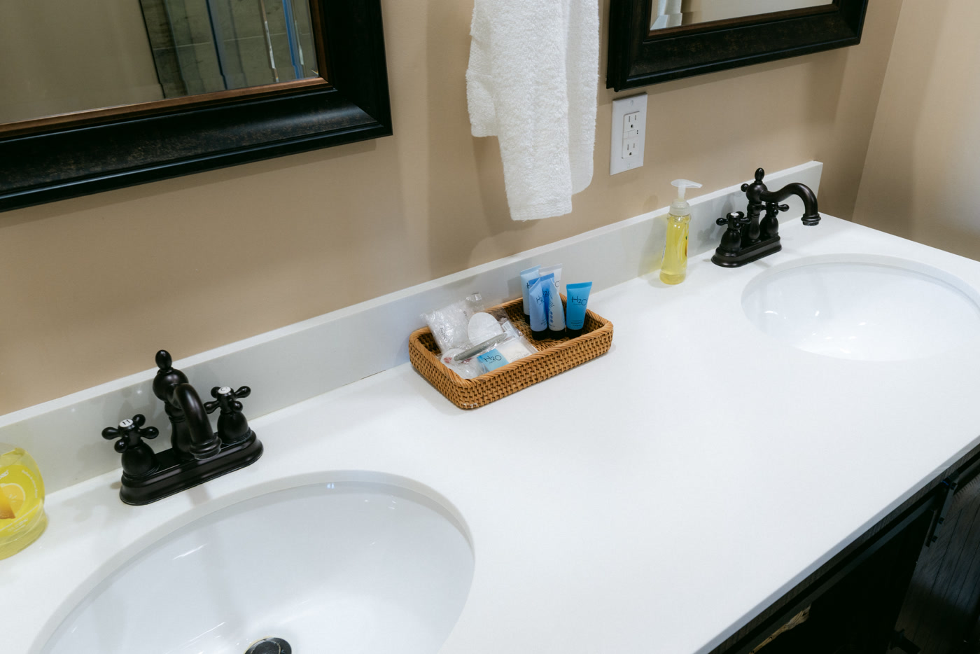 The Elk Suite's double vanity at Shire Ridge Lodge near Bragg Creek: two white basins with black faucets, a basket of toiletries between them and dark framed mirrors above