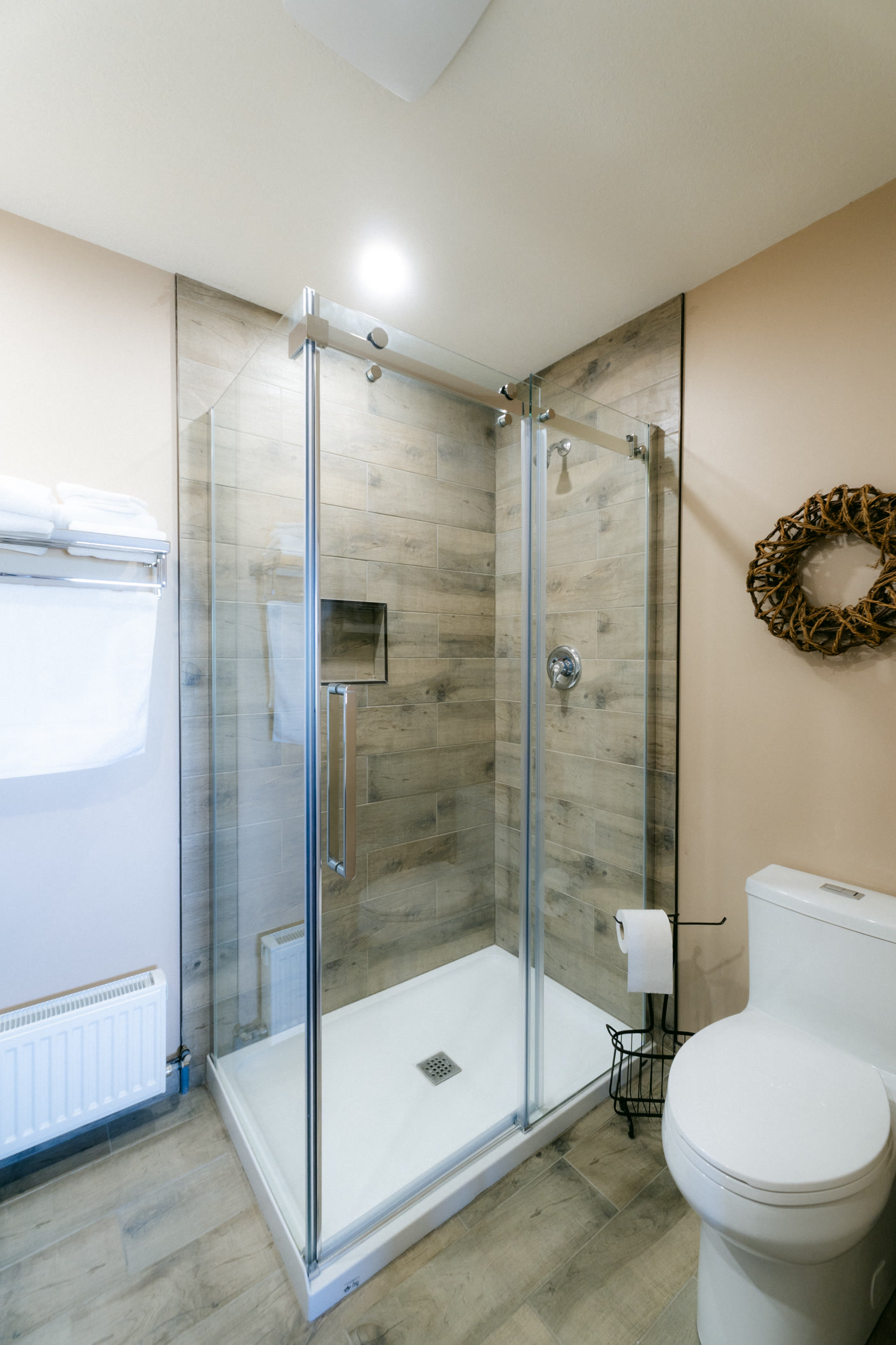 The Elk Suite's walk-in shower at Shire Ridge Lodge near Bragg Creek behind a glass door, wood-look tile on the wall, a white toilet beside it and a woven wreath hung up