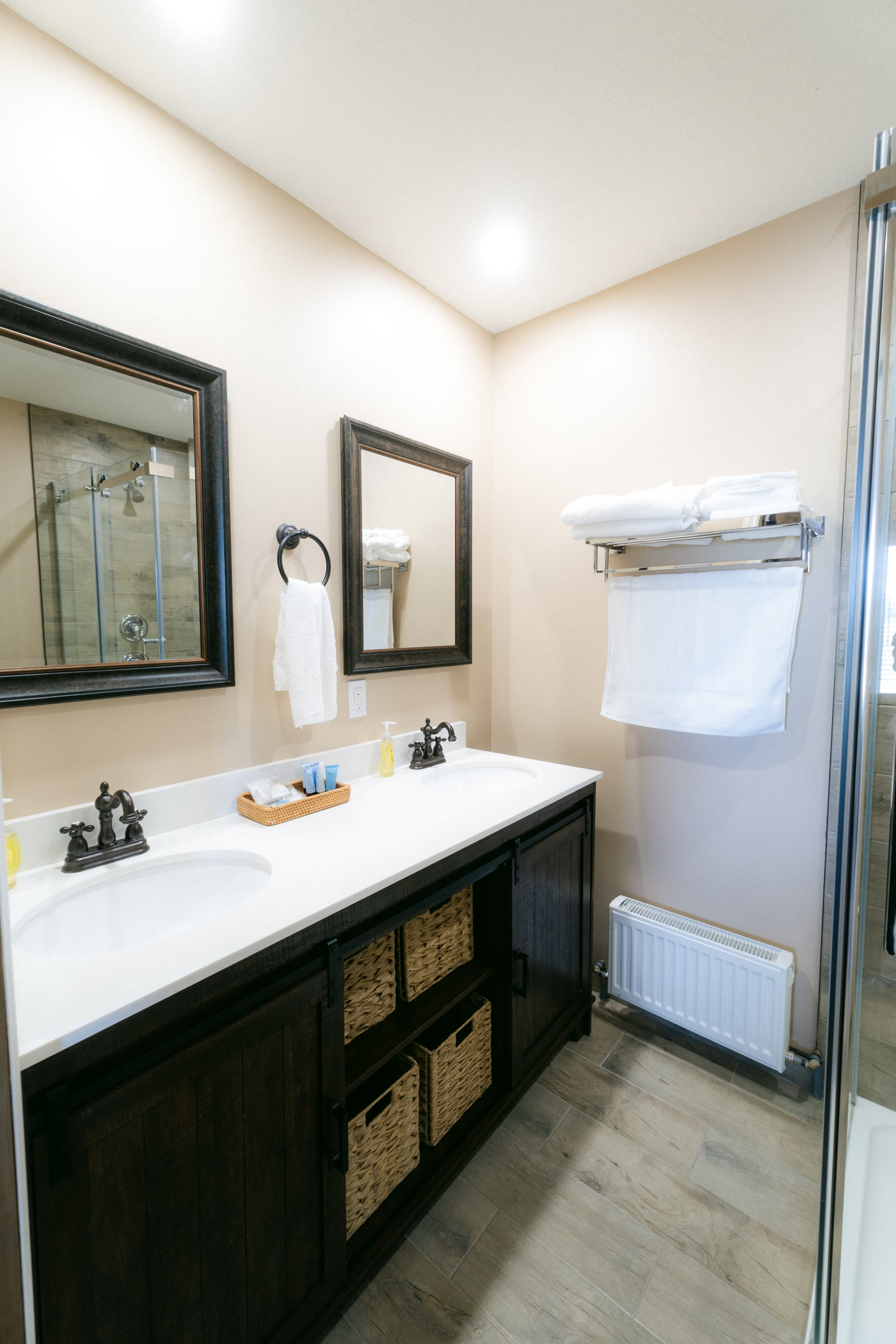 The Elk Suite's double vanity from the side at Shire Ridge Lodge near Bragg Creek: dark cabinets with woven baskets, white towels on a rack, and two framed mirrors on the wall