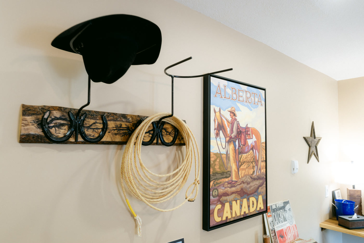 A black cowboy hat and a coiled lariat on horseshoe hooks along a barn-board rack in the Stampede Room at Shire Ridge Lodge near Bragg Creek, beside an Alberta Canada poster of a cowboy and his horse