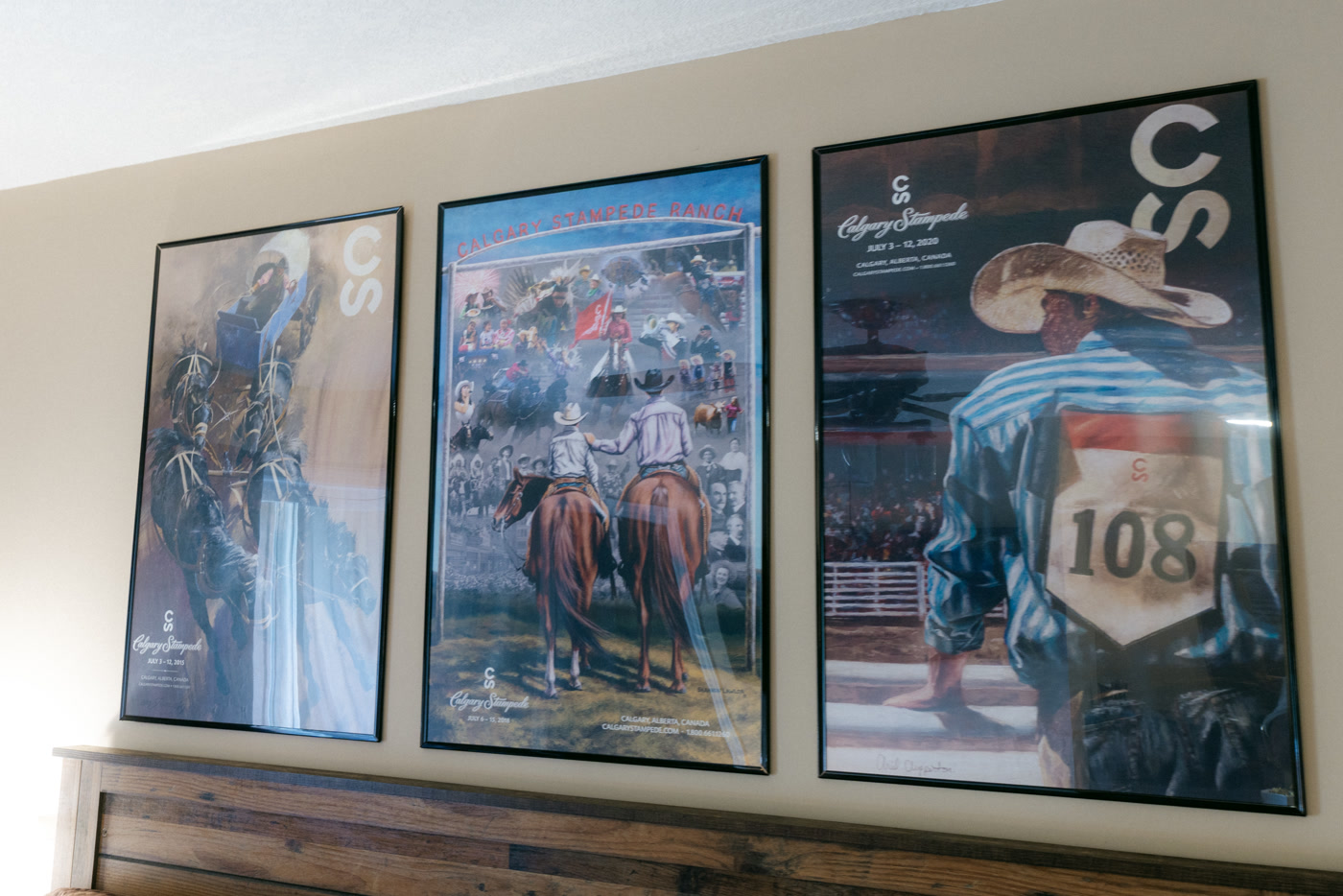 Three framed Calgary Stampede posters above the headboard in the Stampede Room at Shire Ridge Lodge near Bragg Creek: a chuckwagon driver and his team, two riders under a Calgary Stampede Ranch sign, and a cowboy wearing number 108