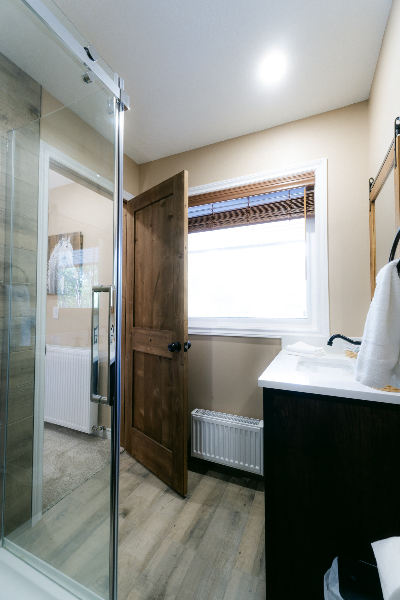 Through the open glass shower door in the Stampede Room's bathroom at Shire Ridge Lodge near Bragg Creek: a window above a radiator, a wooden door and the dark vanity