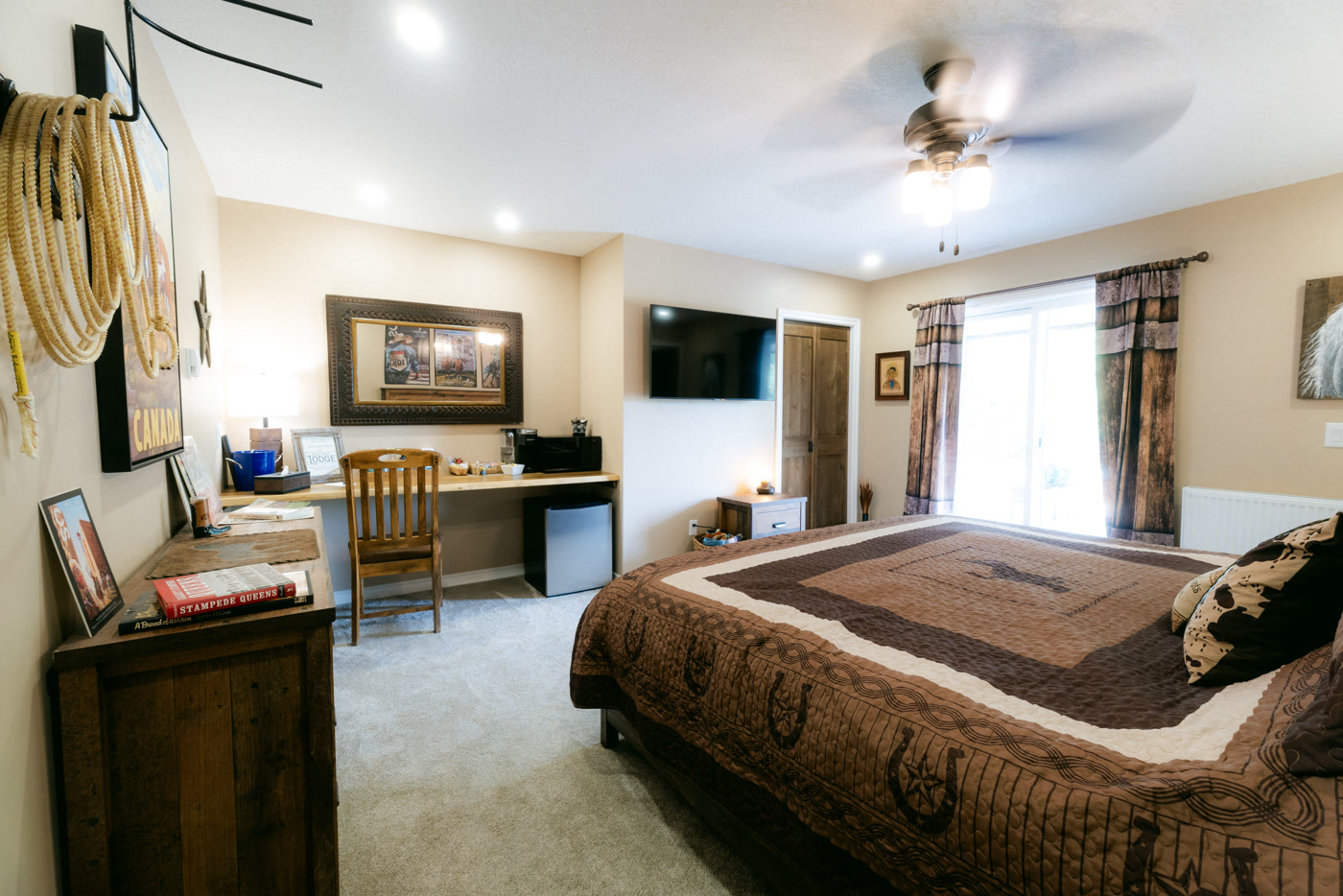 The Stampede Room at Shire Ridge Lodge near Bragg Creek: a king bed under a brown horseshoe-pattern quilt, a coiled lariat on the wall, a desk nook, a ceiling fan and a window