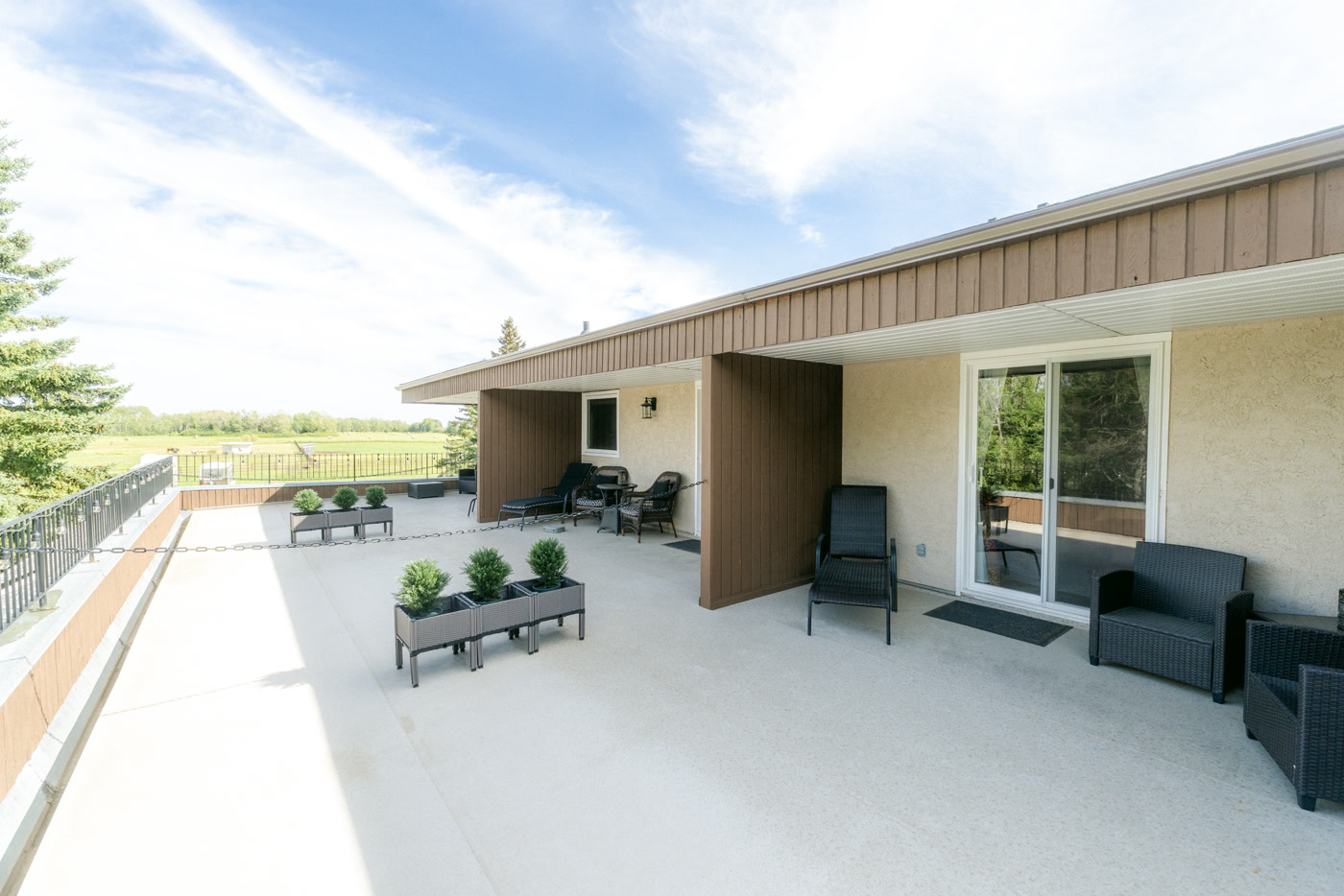 The long balcony along Shire Ridge Lodge near Bragg Creek: a pale deck with planters of boxwood, a covered alcove of lounge chairs, glass railing, and green fields beyond it