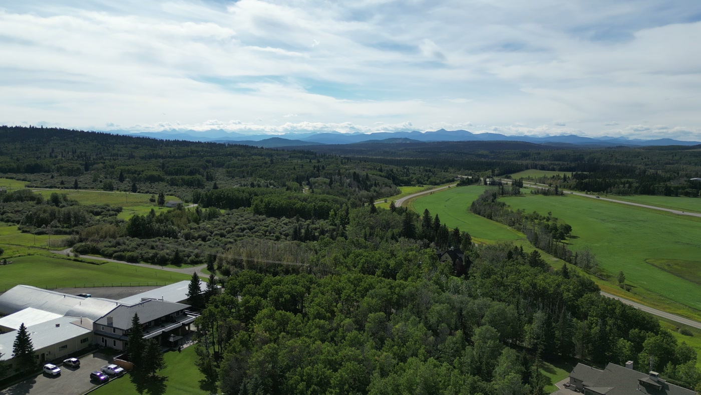 From above Shire Ridge Ranch near Bragg Creek: the lodge and its white barns at the lower edge, green fields and forest running out to a line of mountains under grey-white cloud