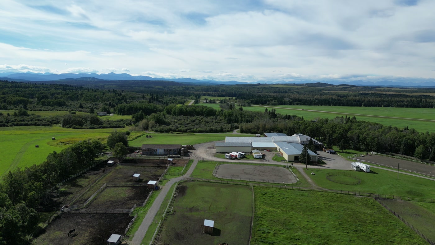 The paddocks, shelters, sand rings and white arena buildings of Shire Ridge Ranch near Bragg Creek from above, fields and forest beyond them, and the mountains along the horizon